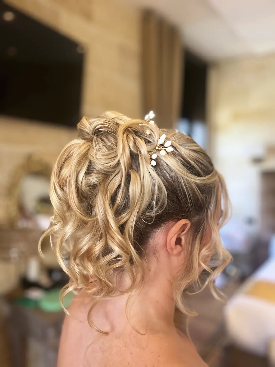 Chignon haut bouclé et volumineux sur cheveux blonds avec bijou de tête perlé, mèches libres sur la nuque