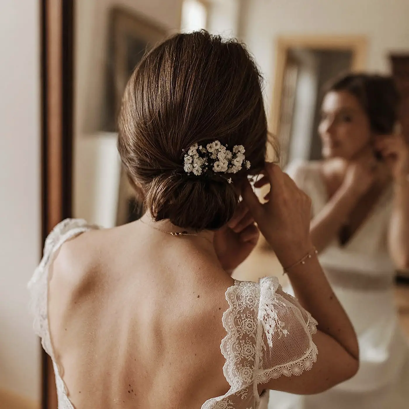 Chignon bas lisse sur cheveux bruns avec bijou perlé, mariée en robe de dentelle dos nu devant le miroir