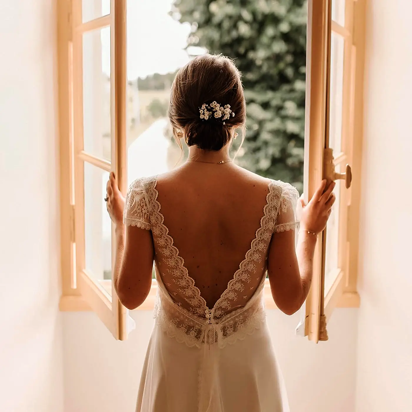 Mariée de dos devant une fenêtre ouverte, chignon bas lisse fleuri et robe de dentelle